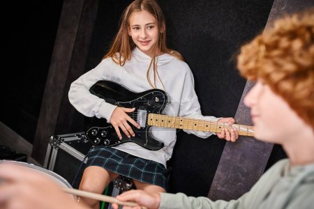 Photo for Focus on jolly pretty teenage girl holding her guitar and looking at her blurred cute drummer - Royalty Free Image