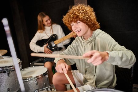 Photo for Focus on joyful cute teenage boy in casual attire playing drums next to his blurred cute guitarist - Royalty Free Image