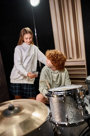 Photo for Cheerful cute teenage band members in casual attires looking at mobile phone near drum set - Royalty Free Image
