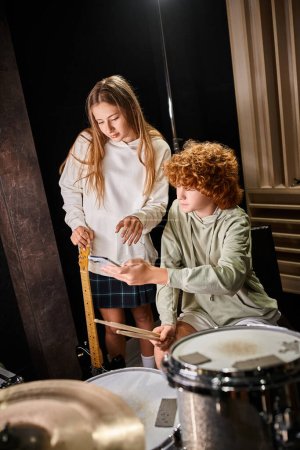 Photo for Focused adorable teenage musicians looking at smartphone next to drum set with guitar in hand - Royalty Free Image