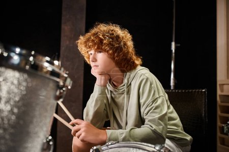 konzentrierter süßer rothaariger Teenager in lässiger Kleidung sitzt vor seinem Schlagzeug-Set