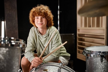 cheerful adorable red haired teenager in casual vivid attire in front of drum set looking at camera