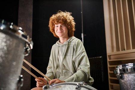 cheerful adorable teenager with red hair in casual outfit in front of his drum set smiling at camera