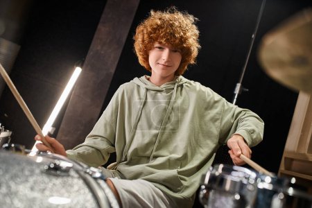 happy cute red haired teenage boy in everyday outfit playing his drums and looking at camera