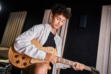 konzentrierter talentierter, entzückender Teenager im lässigen Outfit, der im Studio seine Gitarre spielt