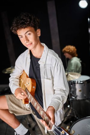 Konzentrierter Teenager in lässiger Kleidung, der in der Nähe seines verschwommenen rothaarigen Schlagzeugers Gitarre spielt