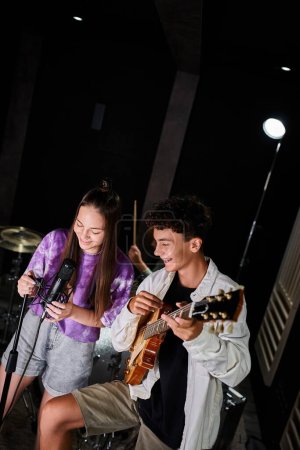 happy talented teenage girl in vivid attire singing into microphone next to her guitarist in studio