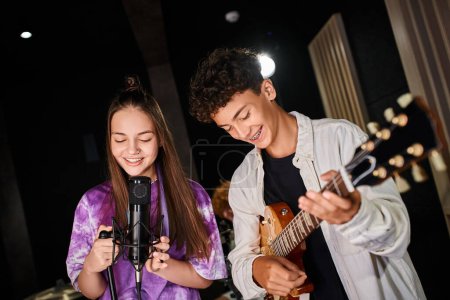 Photo pour Joyeuse adolescente mignonne en tenue vibrante chantant dans le microphone à côté de son guitariste - image libre de droit