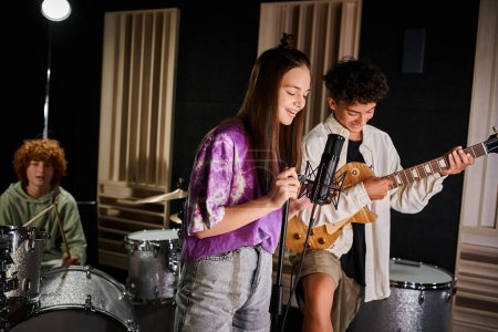 focus on jolly teenage boy and girl singing and playing guitar next to their blurred drummer