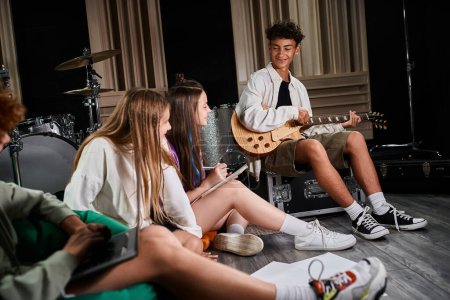 Fokus auf glücklichen Teenie-Jungen in lässiger Kleidung mit Gitarre und Blick auf seine niedlichen Bandmitglieder