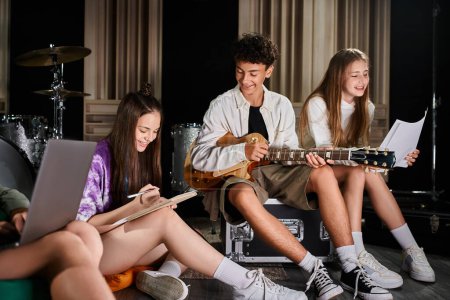 fröhliche, süße Teenager-Bandmitglieder sitzen mit Gitarre und Laptop im Studio und schreiben Texte