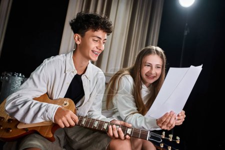 cheerful teenage boy with braces and blonde girl playing guitar and looking at lyrics in studio