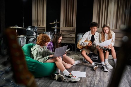 cheerful cute teenage friends sitting on floor with laptop and guitar looking at lyrics in studio