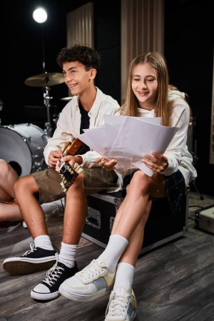 lustiges Teenager-Mädchen, das neben ihrem Freund mit Hosenträgern, die Gitarre spielen, Texte anschaut, Musikgruppe