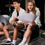 jolly teenage girl looking at lyrics next to her friend with braces playing guitar, musical group