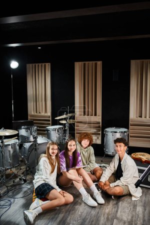 talentueux adolescents joyeux assis en studio avec des paroles et regardant la caméra, groupe musical
