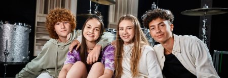 Foto de Adolescentes felices en trajes casuales sentados en el estudio y mirando a la cámara, grupo musical, pancarta - Imagen libre de derechos