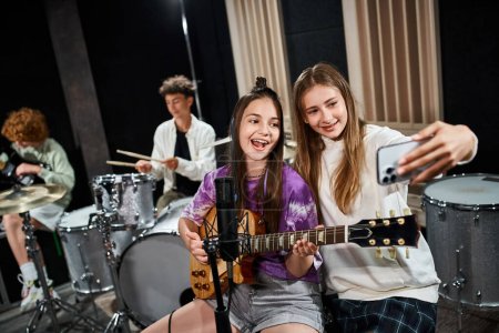 Photo for Focus on jolly teenage girls taking selfie and singing with blurred friends playing instruments - Royalty Free Image