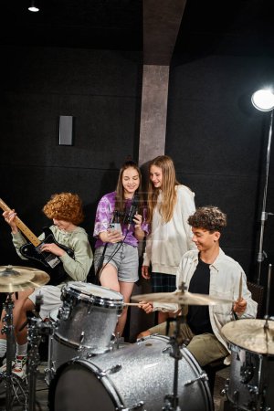 Foto de Alegre adolescentes cantando mientras los niños tocando su batería y guitarra en el estudio, grupo musical - Imagen libre de derechos