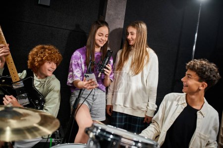 Foto de Alegre lindo adolescente niñas cantando y sonriendo mientras niños jugando guitarra y batería en estudio - Imagen libre de derechos