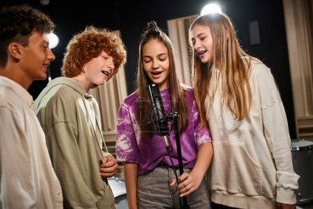 Foto de Alegre lindo adolescentes en casual trajes cantando juntos y sonriendo felizmente en estudio, grupo musical - Imagen libre de derechos