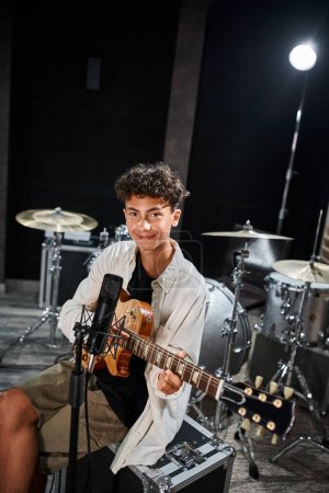 Foto de Alegre talentoso adolescente en traje casual tocando su guitarra y mirando a la cámara en el estudio - Imagen libre de derechos