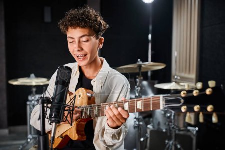 Foto de Talentoso lindo adolescente en traje casual tocando la guitarra y cantando canción en el micrófono en el estudio - Imagen libre de derechos