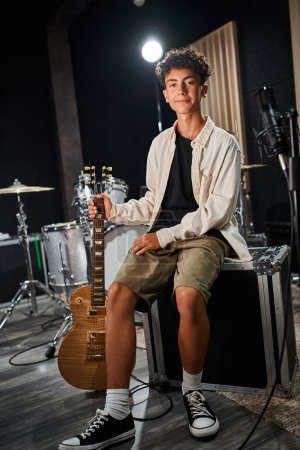Foto de Guapo talentoso adolescente en traje casual sentado y sosteniendo la guitarra y mirando a la cámara - Imagen libre de derechos