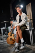 cheerful good looking teenage boy in casual attire holding guitar and smiling at camera in studio hoodie #687123954