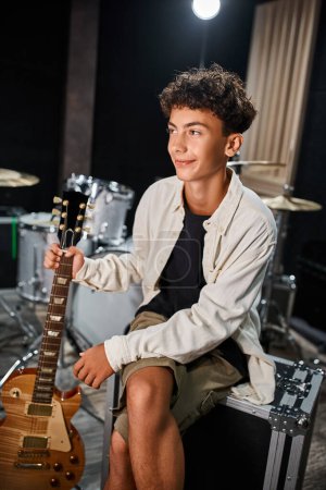 good looking jolly teenage musician in casual attire holding his guitar and looking away in studio