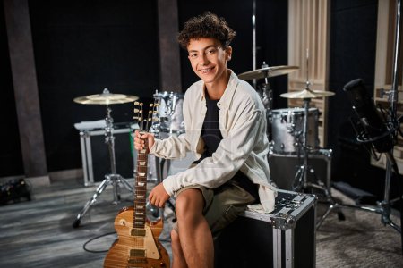 Foto de Guapo talentoso adolescente en traje casual con frenos sosteniendo la guitarra y sonriendo a la cámara en el estudio - Imagen libre de derechos