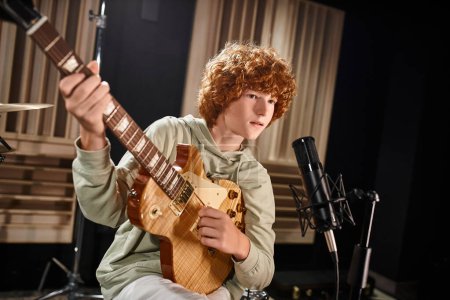 red haired adorable teenage boy in casual attire playing guitar and singing into microphone