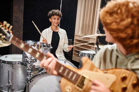 focus on jolly teenage boy playing drums and looking at his red haired blurred friend, musical group
