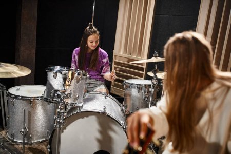 Photo for Jolly adorable teenage girl playing drums while her blonde friend looking at her, musical group - Royalty Free Image