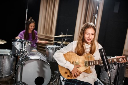 Photo pour Se concentrer sur une jolie adolescente guitariste blonde jouant à côté de son ami flou jouant de la batterie en studio - image libre de droit