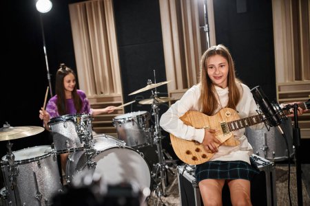 Photo for Jolly talented adorable teenage girls playing guitar and drums while in studio, musical group - Royalty Free Image
