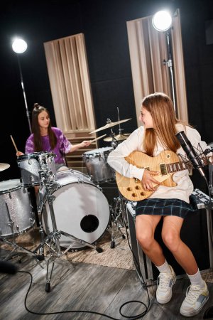 Photo for Pretty blonde teenage girl with guitar looking at her brunette friend playing drums in studio - Royalty Free Image