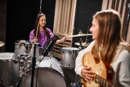 Photo for Focus on pretty brunette teenage drummer next to blonde blurred girl playing guitar in studio - Royalty Free Image