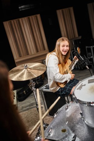 Photo for Jolly teenage girl in casual outfit playing guitar and looking at her friend playing drums - Royalty Free Image