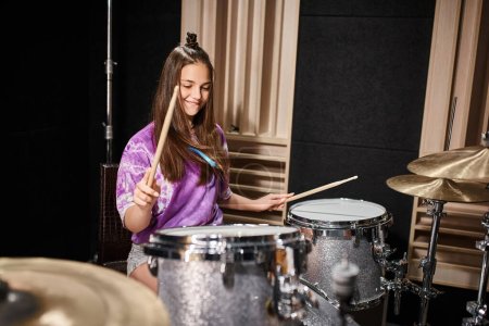 Photo pour Belle brunette adolescente en tenue décontractée vibrante jouer de la batterie tandis que dans le studio de musique - image libre de droit