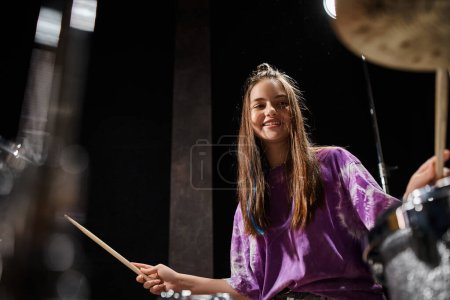 jolly adorable brunette teenage girl in vibrant casual attire playing drums while in studio