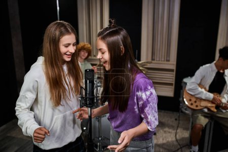 Photo for Pretty teenage girls singing into microphone while boys playing drums and guitar, musical group - Royalty Free Image