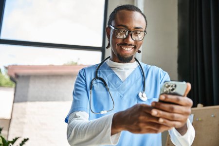 schöner fröhlicher afrikanisch-amerikanischer Arzt mit Stethoskop, der auf sein Handy schaut, Telemedizin