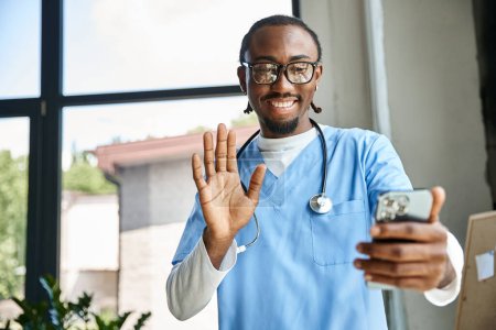 fröhlicher junger afrikanisch-amerikanischer Arzt mit Stethoskop, der seinem Handy zuwinkt, Telemedizin