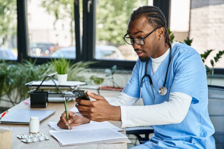 médico afroamericano concentrado con gafas tomando notas y consultando por teléfono, telesalud
