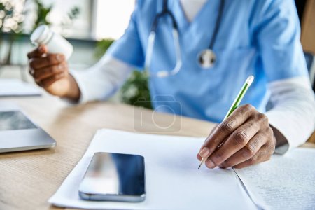 Photo for Cropped view of african american doctor with stethoscope taking notes while working, phone on table - Royalty Free Image