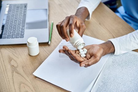 vista recortada de los jóvenes afroamericanos médico profesional verter píldoras en la mano junto a la computadora portátil