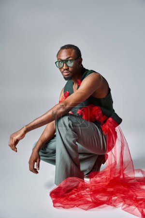 good looking african american man in stylish outfit squatting on gray backdrop, fashion concept