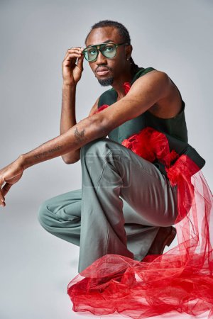 handsome african american man in stylish outfit squatting on gray backdrop, fashion concept