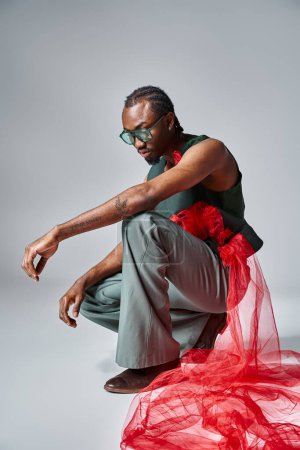 good looking african american man in trendy attire squatting on gray backdrop, fashion concept